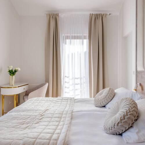 Jasny pokój hotelowy z dużym łóżkiem, pikowaną narzutą, ozdobnymi okrągłymi poduszkami, drewnianym zagłówkiem, beżowymi zasłonami i toaletką z kwiatami.