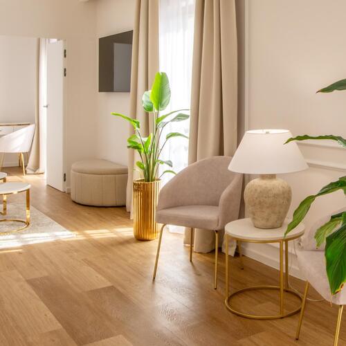Przytulny salon apartamentu z aksamitnymi fotelami, złotymi stolikami, lampą stołową, dużymi roślinami i jasnymi zasłonami