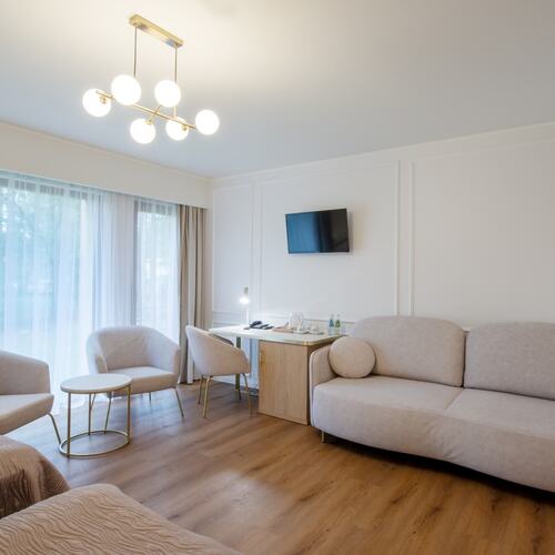 Jasny apartament z beżową sofą i okrągłymi fotelami, stolikiem, ściennym telewizorem, biurkiem z czajnikiem i dużym oknem z firaną
