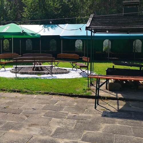 Zewnętrzny ogród hotelowy z ławkami wokół paleniska, zielonymi parasolami, zadaszonymi namiotami, grillem i wiszącymi lampkami.