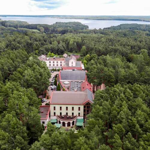 Hotel pośród gęstego sosnowego lasu, z tarasami i parasolami, parkingiem, pokojami z widokiem na oddalone jezioro.