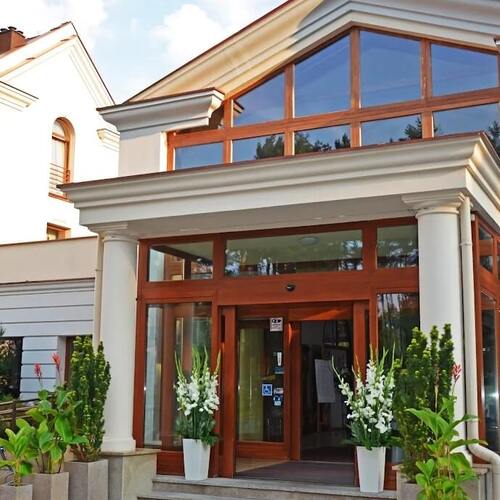 Eleganckie wejście hotelu z kolumnami, dużą przeszkloną fasadą i drewnianymi framugami, kwiaty w donicach, znak dostępności.
