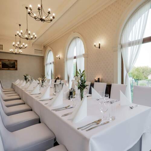 Elegancka sala bankietowa z długim białym stołem, staranną zastawą, świeżymi kwiatami, żyrandolami i łukowymi oknami.