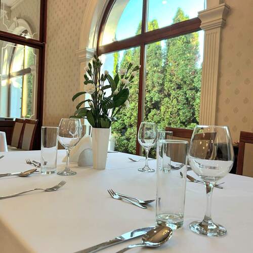 Elegancka sala restauracyjna z białym obrusem, szkłem i sztućcami, wazonem z białymi kwiatami i łukowym oknem na zieleń.
