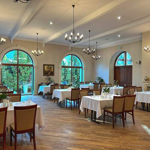 Elegancka jadalnia hotelowa z białymi obrusami, nakrytymi stołami, żyrandolami, dębową podłogą i dużymi półokrągłymi oknami na ogród