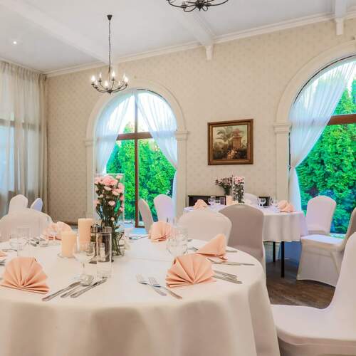 Elegancka sala bankietowa z okrągłymi stołami w białych obrusach, różowymi serwetkami, kwiatowymi dekoracjami, łukowymi oknami i żyrandolem