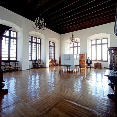 Przestronna sala z lśniącą drewnianą podłogą, wysokimi arkadowymi oknami, żyrandolami i antycznymi meblami, zwiedzający.
