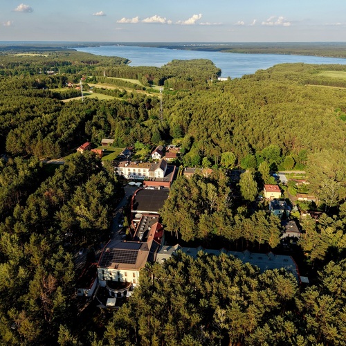 Kompleks hotelowy w gęstym sosnowym lesie, z tarasami i panelami słonecznymi na dachu, w tle rozległe jezioro