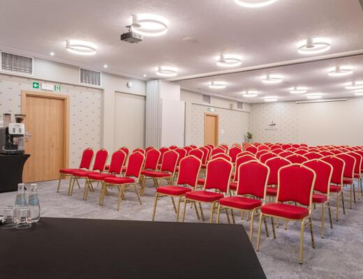Sala konferencyjna w układzie teatralnym z czerwonymi krzesłami, projektorem sufitowym, nowoczesnym oświetleniem i stacją kawową