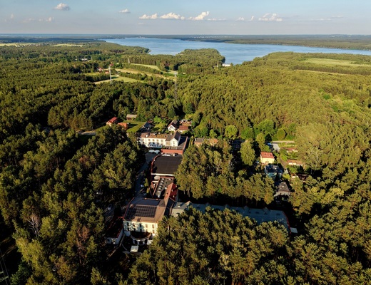 Kompleks hotelowy w gęstym sosnowym lesie, z tarasami i panelami słonecznymi na dachu, w tle rozległe jezioro