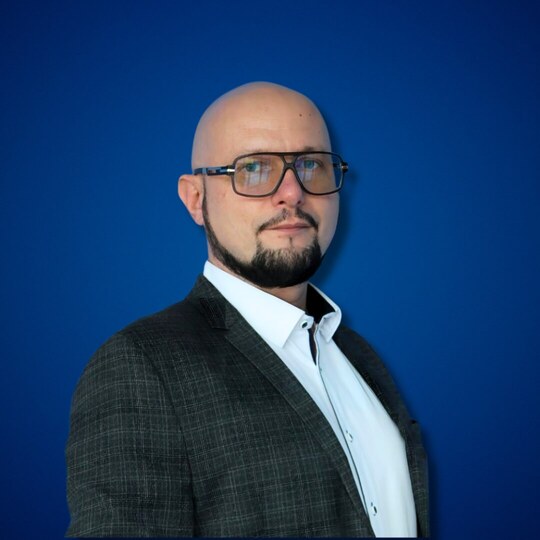 Mężczyzna w marynarce i białej koszuli, w dużych okularach i krótką przyciętą brodą, na gładkim niebieskim tle