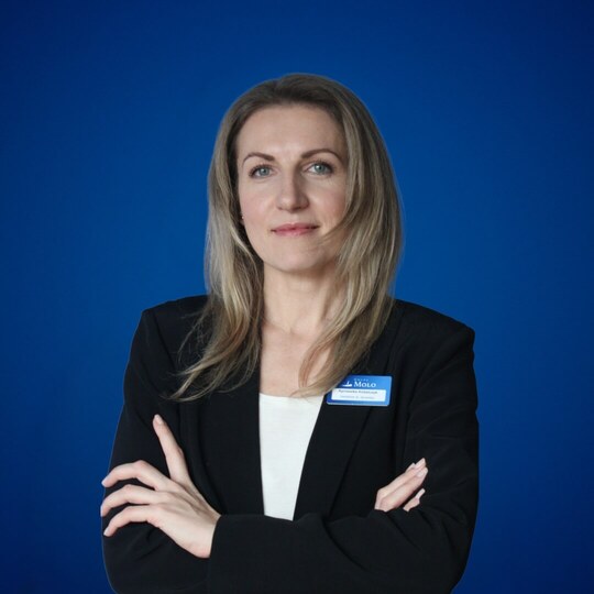 Pracowniczka hotelu w czarnej marynarce i białej bluzce, z plakietką imienną, z założonymi rękami na niebieskim tle.