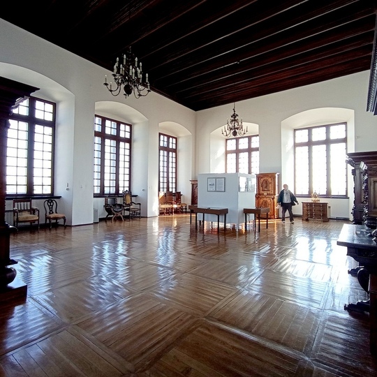 Przestronna sala z lśniącą drewnianą podłogą, wysokimi arkadowymi oknami, żyrandolami i antycznymi meblami, zwiedzający.