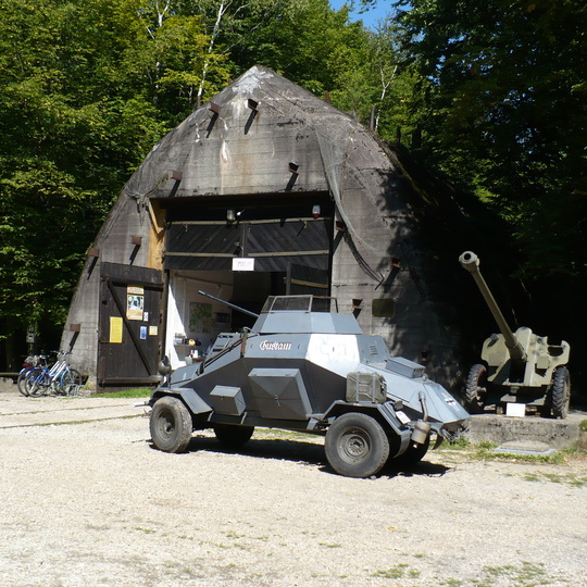 Betonowy schron wśród drzew z otwartymi drewnianymi drzwiami, przed nim szary opancerzony samochód i armata, rowery przy wejściu.