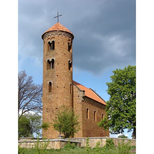 Okrągła romańska wieża kościelna z cegły i stożkowym czerwonym dachem, otoczona kamiennym murem i zielonymi drzewami