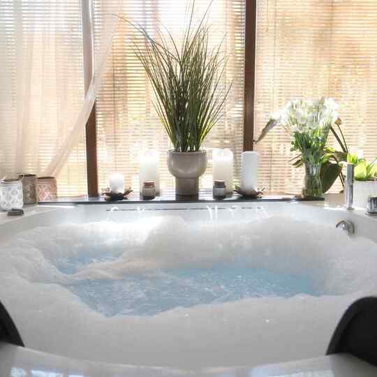 Jacuzzi z bąbelkami w strefie spa, świece, doniczkowe rośliny i naturalne światło przez żaluzje.