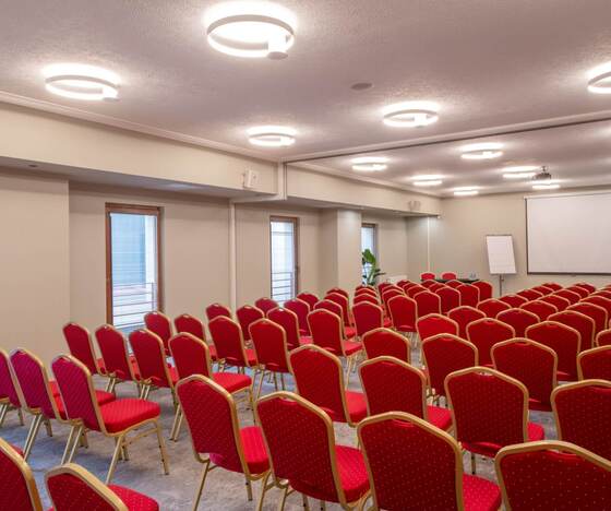 Sala konferencyjna z rzędami czerwonych krzeseł, ekranem i projektorem, flipchartem i jasnym okrągłym oświetleniem