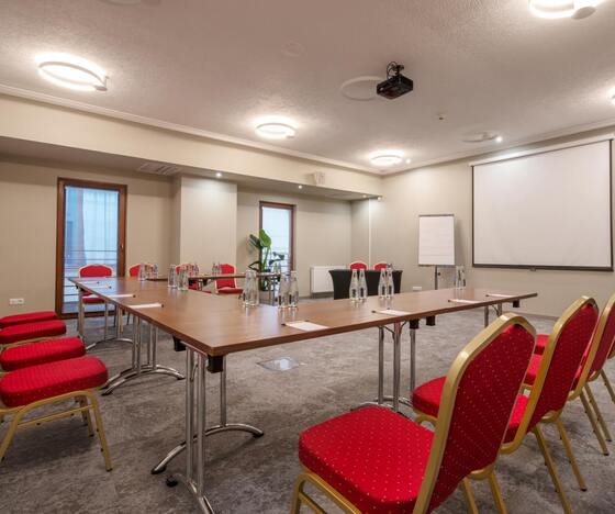 Sala konferencyjna w układzie podkowy z czerwonymi krzesłami, stołami, projektorem, ekranem, flipchartem i butelkami wody.