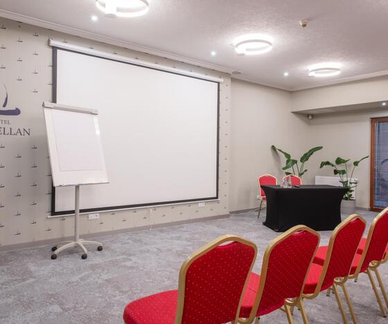 Sala konferencyjna Hotel Magellan: ekran projekcyjny, flipchart, rzędy czerwonych krzeseł, stół prelegenta i rośliny.