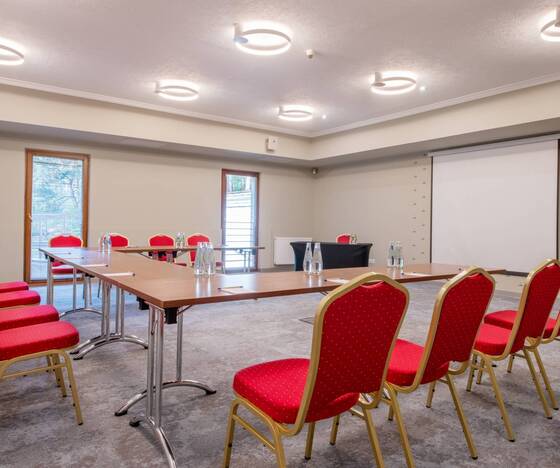 Sala konferencyjna z czerwonymi krzesłami, stołami w literę U, ekranem projekcyjnym, butelkami wody i nowoczesnymi okrągłymi lampami.