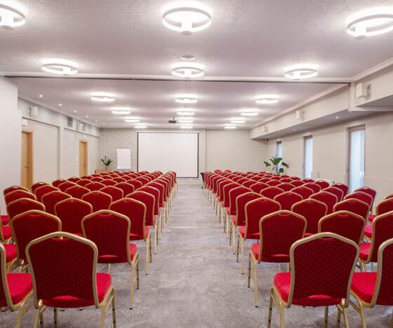 Sala konferencyjna z czerwonymi krzesłami w rzędach, projektorem i ekranem, nowoczesne oświetlenie i neutralne wnętrze