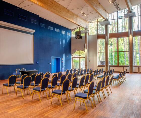 Sala konferencyjna z rzędami krzeseł, ekranem projektora, drewnianymi belkami i dużymi oknami na sosnowy las.