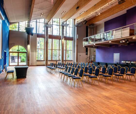 Sala konferencyjna z rzędami krzeseł, wysokimi oknami z widokiem na las, drewnianymi belkami i antresolą.