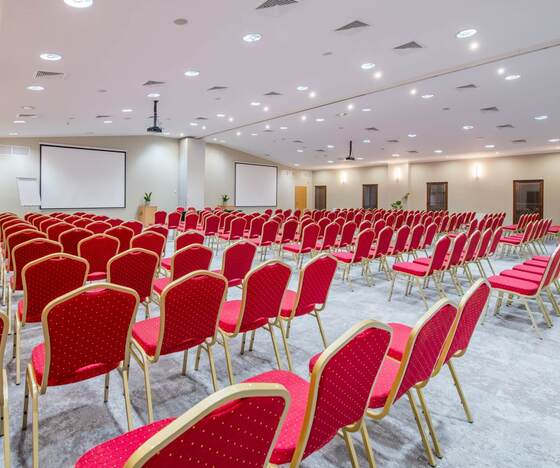 Przestronna sala konferencyjna z rzędami czerwonych krzeseł, dwoma ekranami, projektorem, mównicą i dyskretnym oświetleniem sufitowym