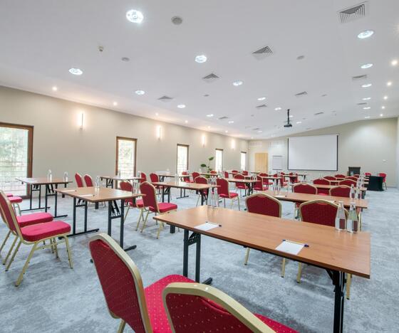 Konferencyjna sala z ustawieniem klasowym, czerwonymi krzesłami, stołami, ekranem projektora, butelkami wody i jasnym oświetleniem