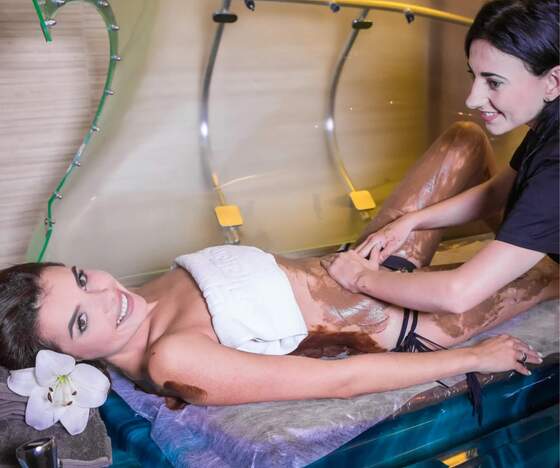 Kobieta podczas zabiegu czekoladowego okładu na leżance, terapeutka masuje nogi; ręcznik, lilia i relaksacyjna kabina spa.