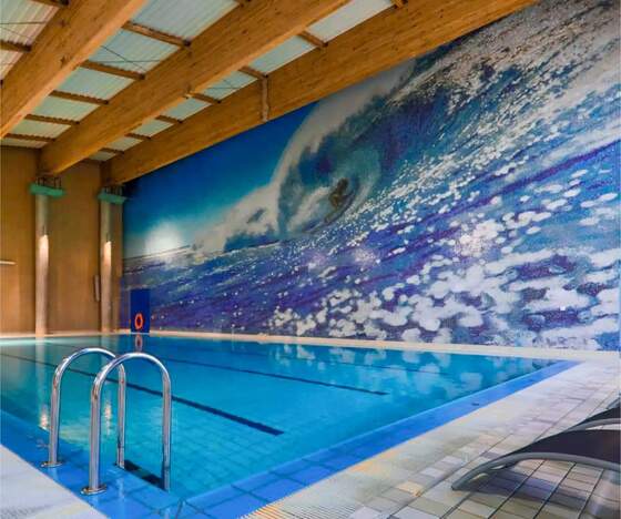 Kryty basen hotelowy z mozaikowym muralem wielkiej fali, drewnianym stropem, metalową drabinką i leżakami