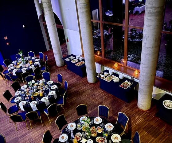 Elegancka sala bankietowa z wysokimi filarami i dużymi oknami, okrągłe stoliki nakryte do kolacji oraz bufet.