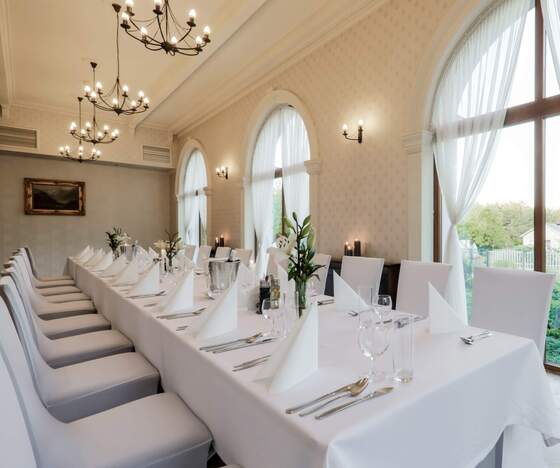 Elegancka sala bankietowa z długim białym stołem, staranną zastawą, świeżymi kwiatami, żyrandolami i łukowymi oknami.