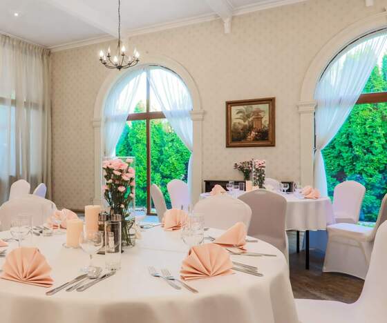 Elegancka sala bankietowa z okrągłymi stołami w białych obrusach, różowymi serwetkami, kwiatowymi dekoracjami, łukowymi oknami i żyrandolem