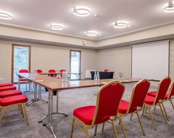 Sala konferencyjna z czerwonymi krzesłami, stołami w literę U, ekranem projekcyjnym, butelkami wody i nowoczesnymi okrągłymi lampami.