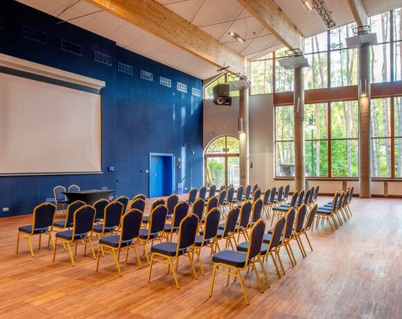 Sala konferencyjna z rzędami krzeseł, ekranem projektora, drewnianymi belkami i dużymi oknami na sosnowy las.