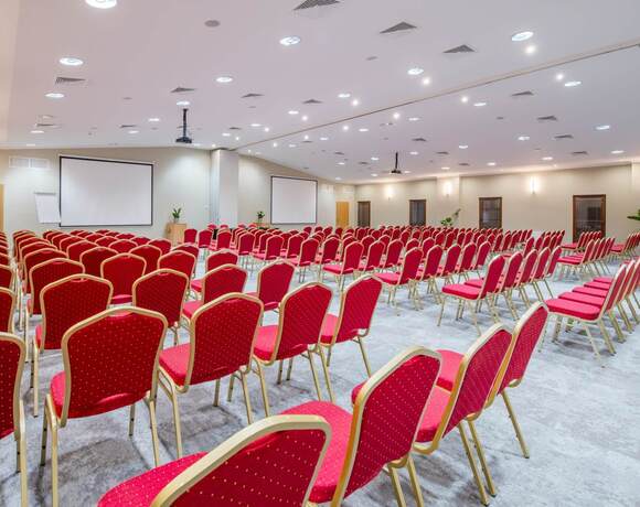 Przestronna sala konferencyjna z rzędami czerwonych krzeseł, dwoma ekranami, projektorem, mównicą i dyskretnym oświetleniem sufitowym