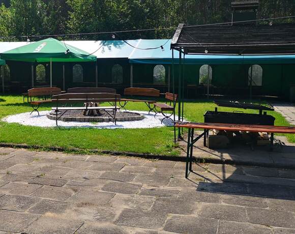 Zewnętrzny ogród hotelowy z ławkami wokół paleniska, zielonymi parasolami, zadaszonymi namiotami, grillem i wiszącymi lampkami.