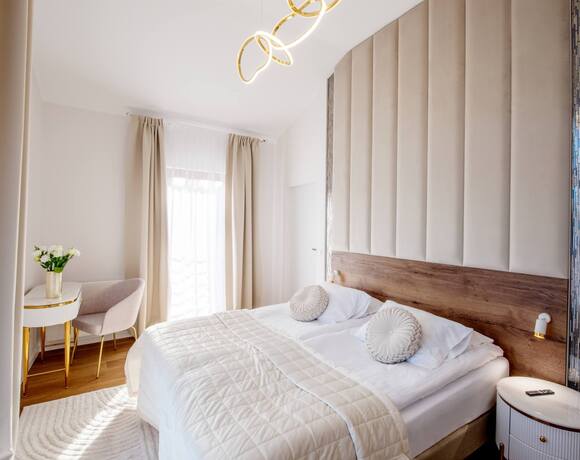 Przytulny pokój hotelowy z dużym łóżkiem, pikowaną narzutą, ozdobnym tapicerowanym zagłówkiem, toaletką ze złotymi nogami i delikatnymi zasłonami.