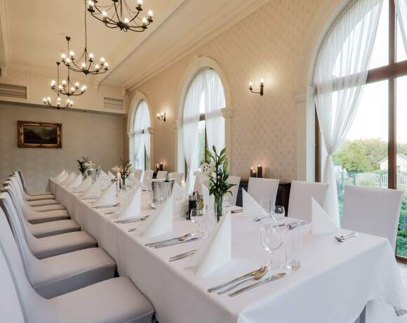 Elegancka sala bankietowa z długim białym stołem, staranną zastawą, świeżymi kwiatami, żyrandolami i łukowymi oknami.