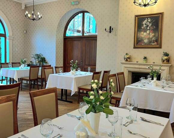 Sala restauracyjna hotelu: białe obrusy, eleganckie nakrycia, świeże kwiaty, drewniane krzesła, żyrandole i kominek.