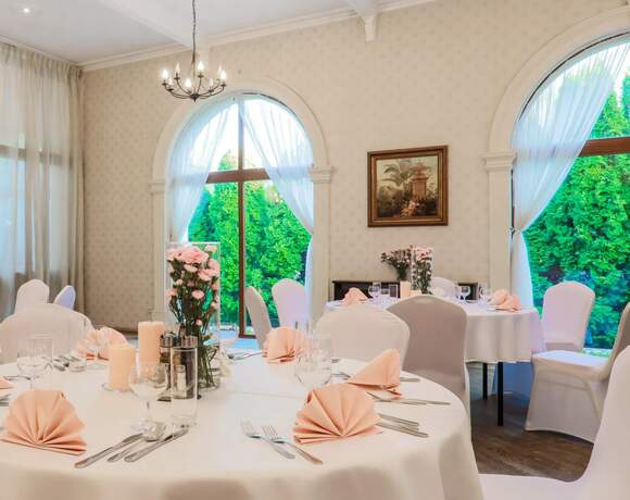 Elegancka sala bankietowa z okrągłymi stołami w białych obrusach, różowymi serwetkami, kwiatowymi dekoracjami, łukowymi oknami i żyrandolem