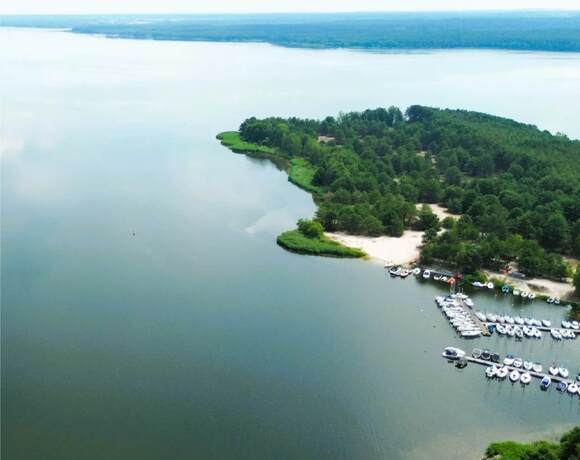 Widok jeziora z zalesionym półwyspem, piaszczystą plażą i małą przystanią z łodziami.