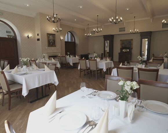 Elegancka sala restauracyjna hotelu — stoły z białymi obrusami i złożonymi serwetkami, kwiaty, łukowe okna i ozdobne żyrandole.