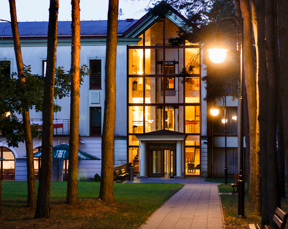 Przeszklone atrium hotelu nocą z ciepłym, oświetlonym lobby; alejka z latarniami i ławkami między sosnami, taras z parasolami