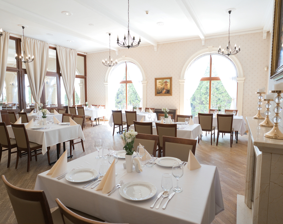 Elegancka restauracja hotelowa z białymi obrusami, żyrandolami, nakrytymi stolikami z talerzami, szkłem i kwiatami, dużymi łukowymi oknami na ogród.