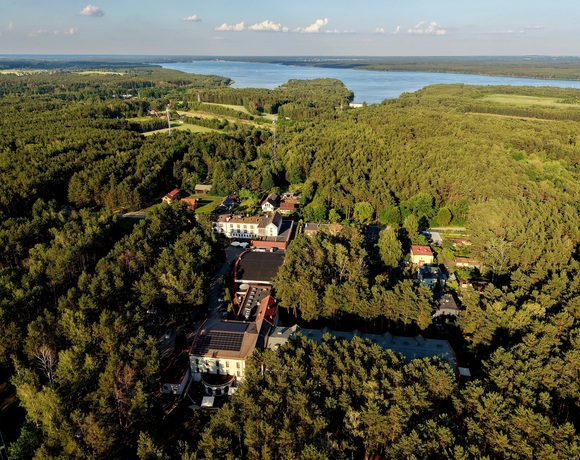Kompleks hotelowy w gęstym sosnowym lesie, z tarasami i panelami słonecznymi na dachu, w tle rozległe jezioro