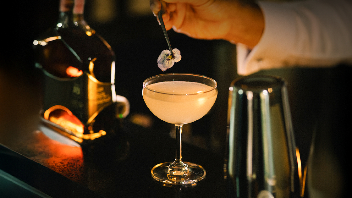 Bartender placing an edible flower on a pale signature cocktail in a coupe glass at a dim, elegant hotel bar, bottle and shaker nearby