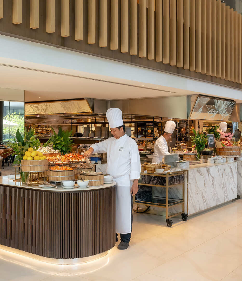 Elegant hotel buffet with marble counters and wooden slat ceiling, chefs arranging seafood and fresh produce at live cooking stations