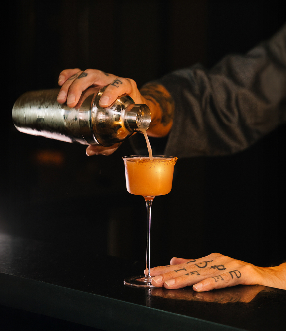Tattooed bartender's hands pouring a shaken orange cocktail from a metal shaker into a rimmed coupe at a dim bar.